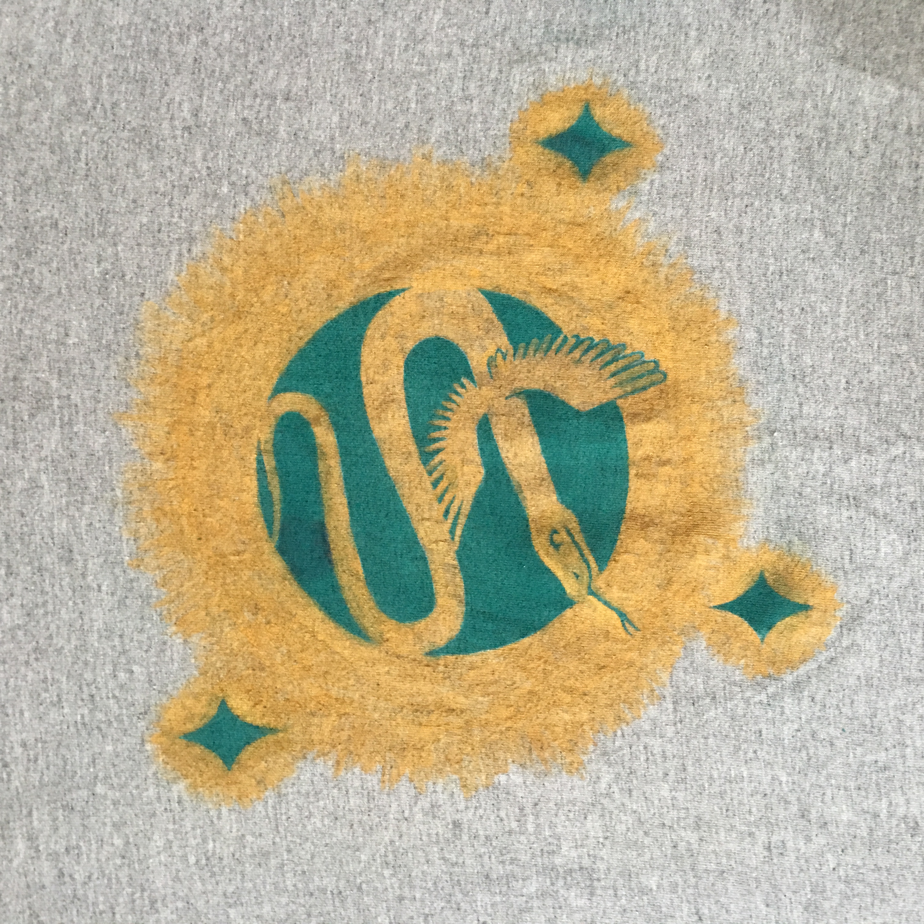 A golden snake with wings surrounded by a hollow golden radial aura with teal stars against a teal background on gray fabric